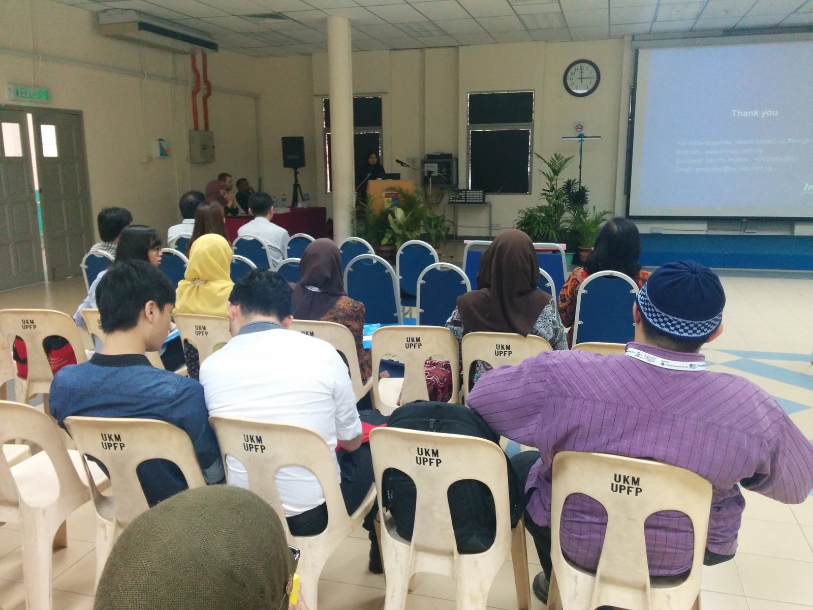Participants seated in rows facing a speaker at the lectern and a projection screen in a hall