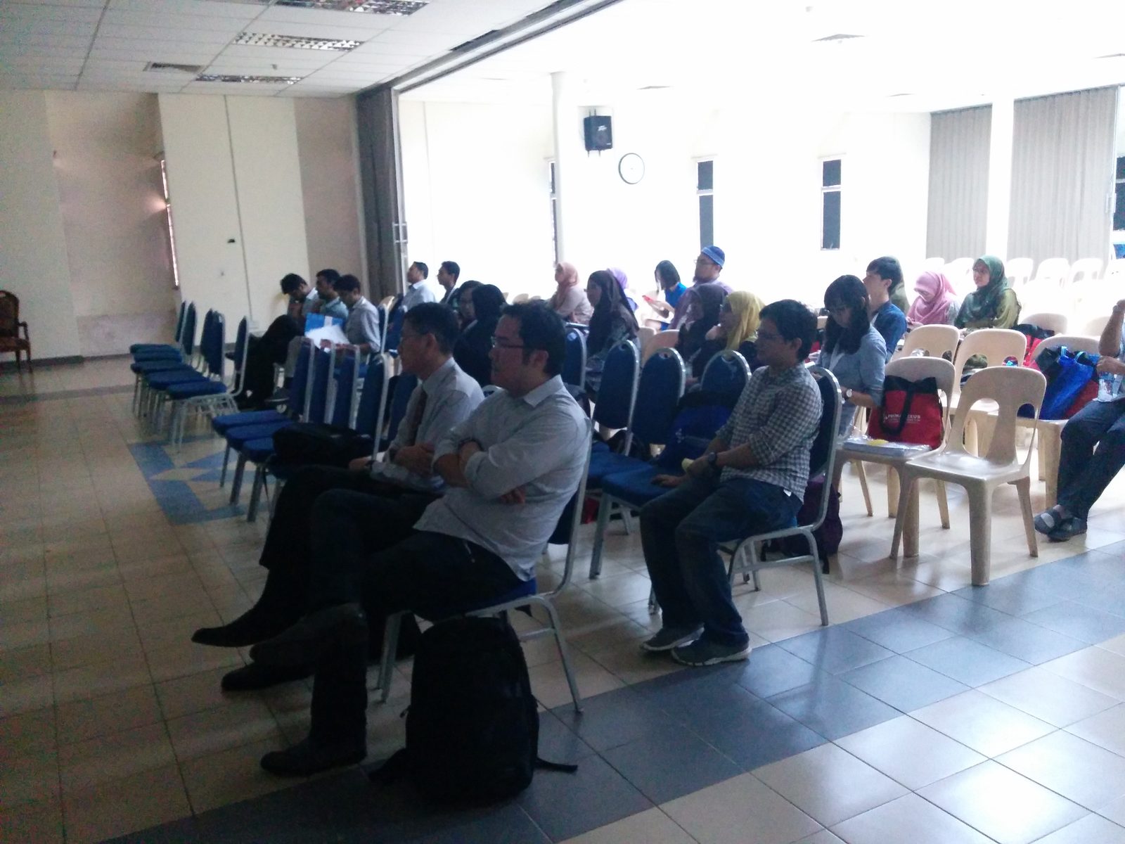 Participants seated in rows of blue and beige chairs in a bright hall during a session