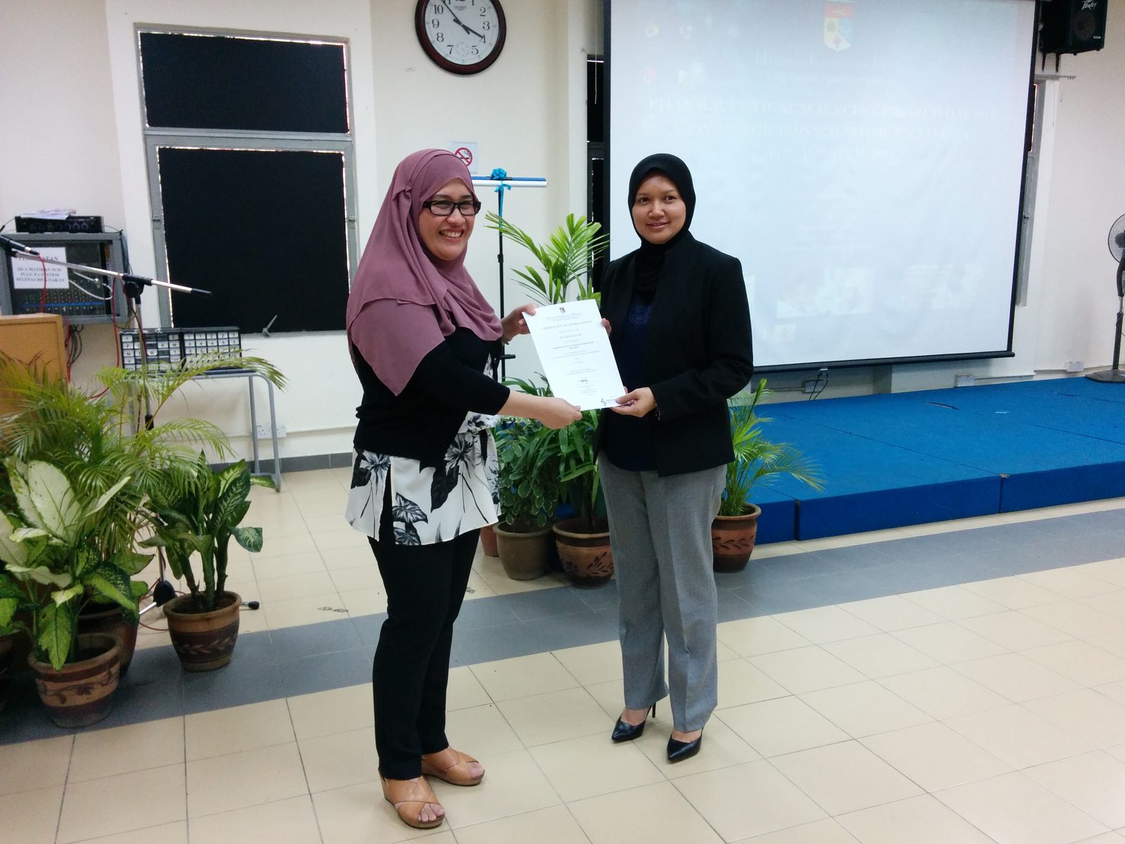 Two women holding a certificate together in front of a projection screen and potted plants