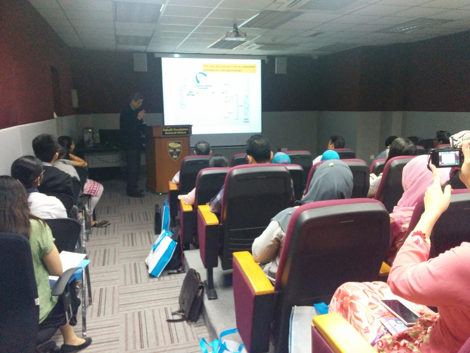A speaker at a lectern presents a slide to a seated audience in a lecture hall