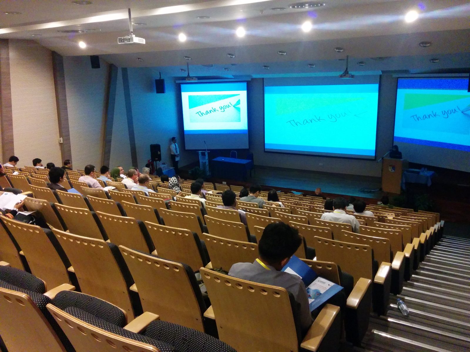 Delegates seated in a tiered lecture hall, facing three large screens
