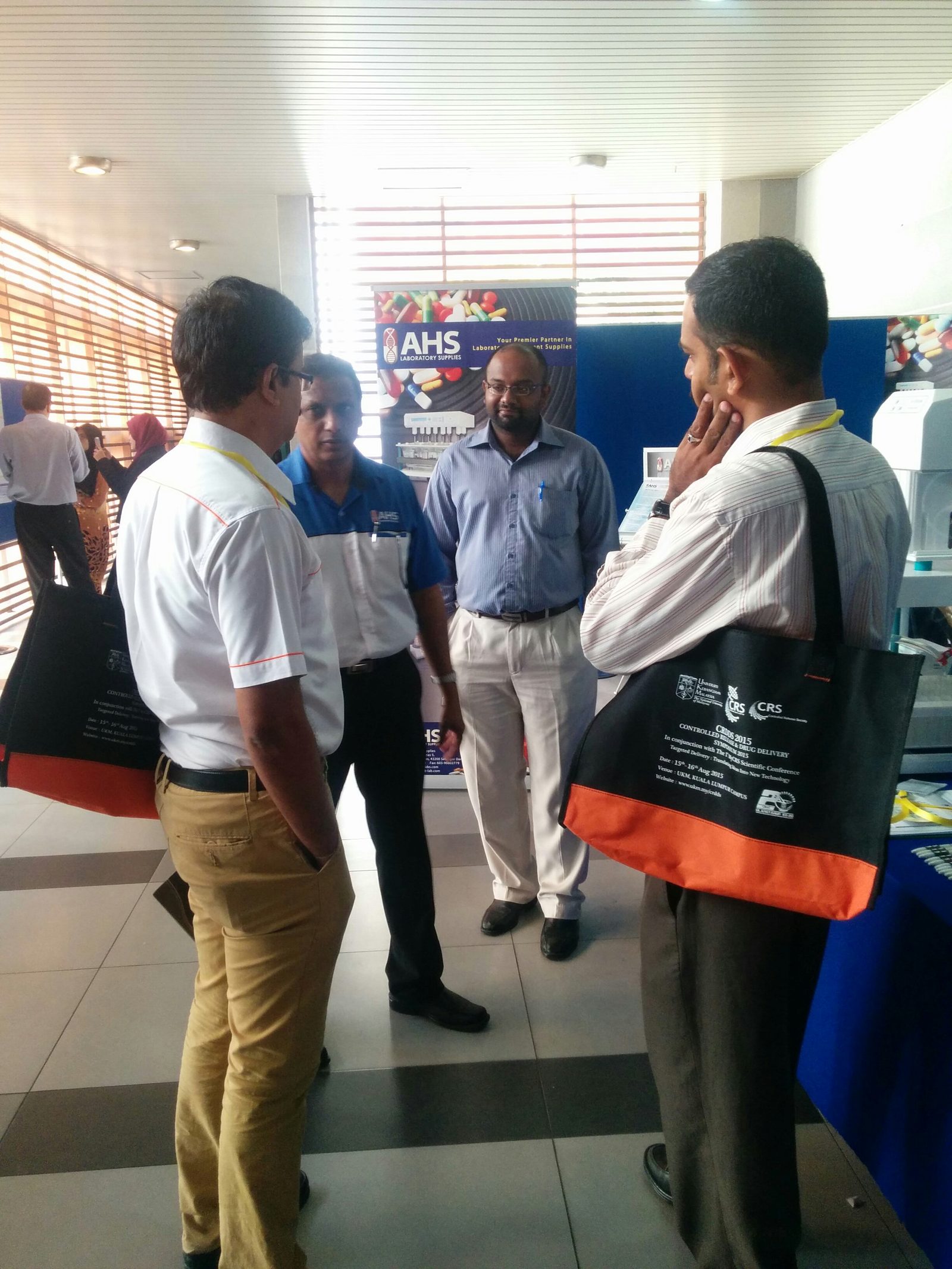Delegates talk at a supplier booth in the exhibition area; two carry the black and orange symposium bags