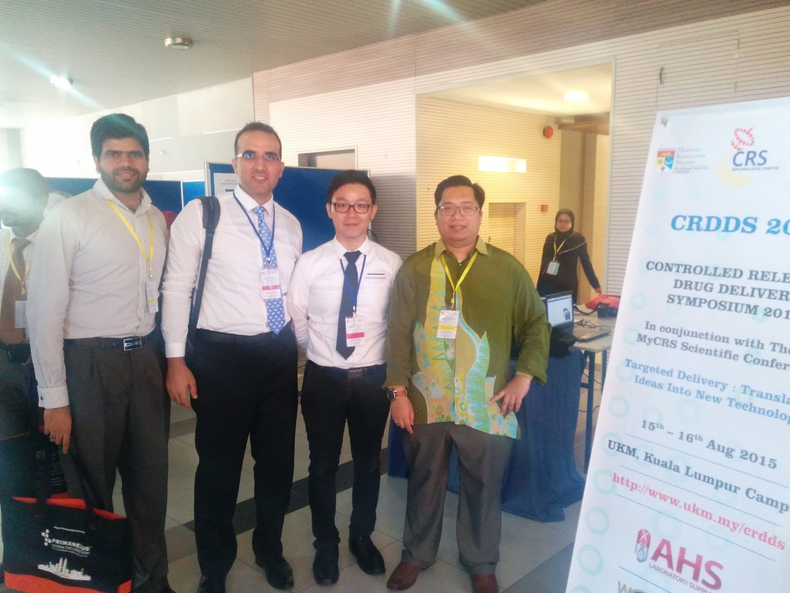 Four delegates stand beside the symposium pull-up banner; one holds a bag printed with the Prima Nexus name