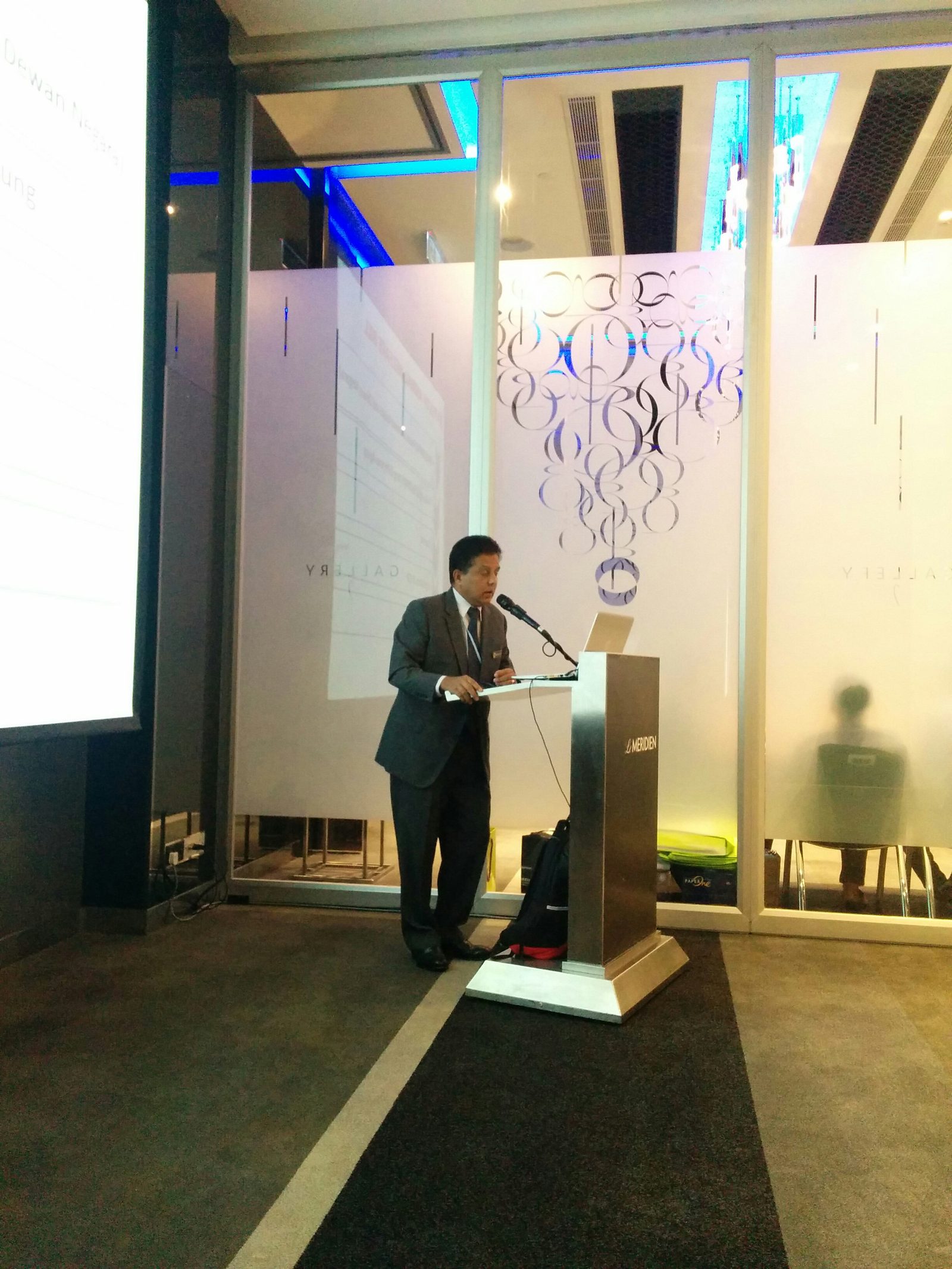 A man in a grey suit speaks at the lectern beside the projection screen