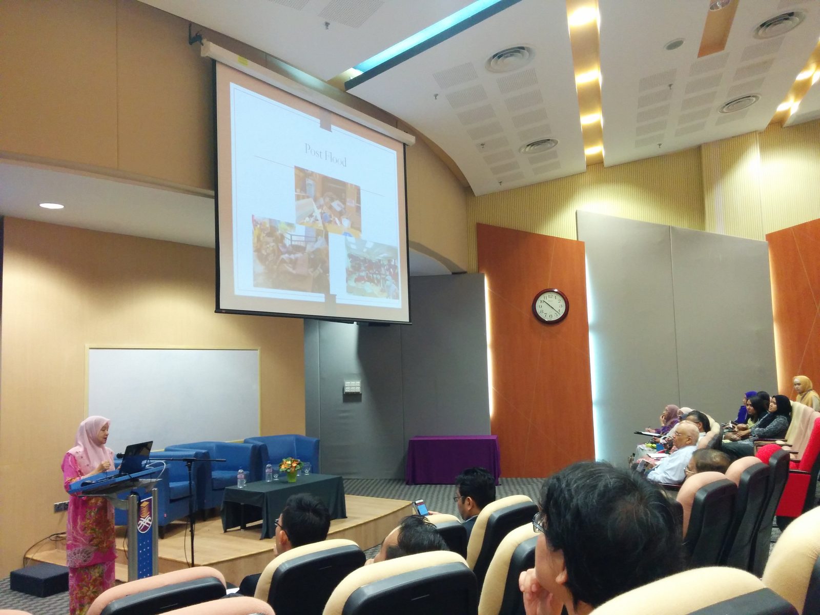 A speaker at the lectern presents a slide titled Post Flood to an audience in a university auditorium