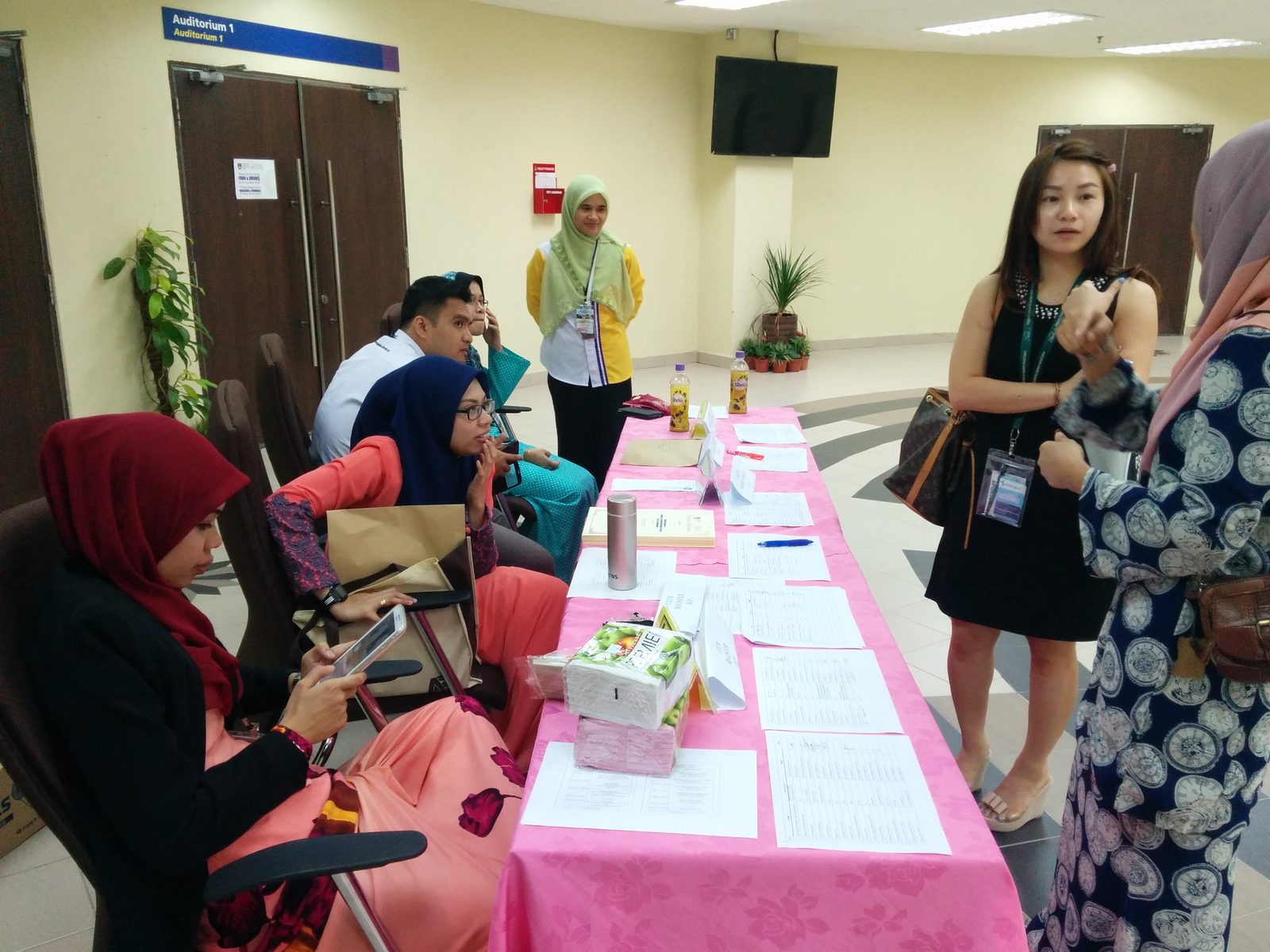 A registration desk with a pink cloth, people seated behind it and attendees wearing lanyards standing nearby