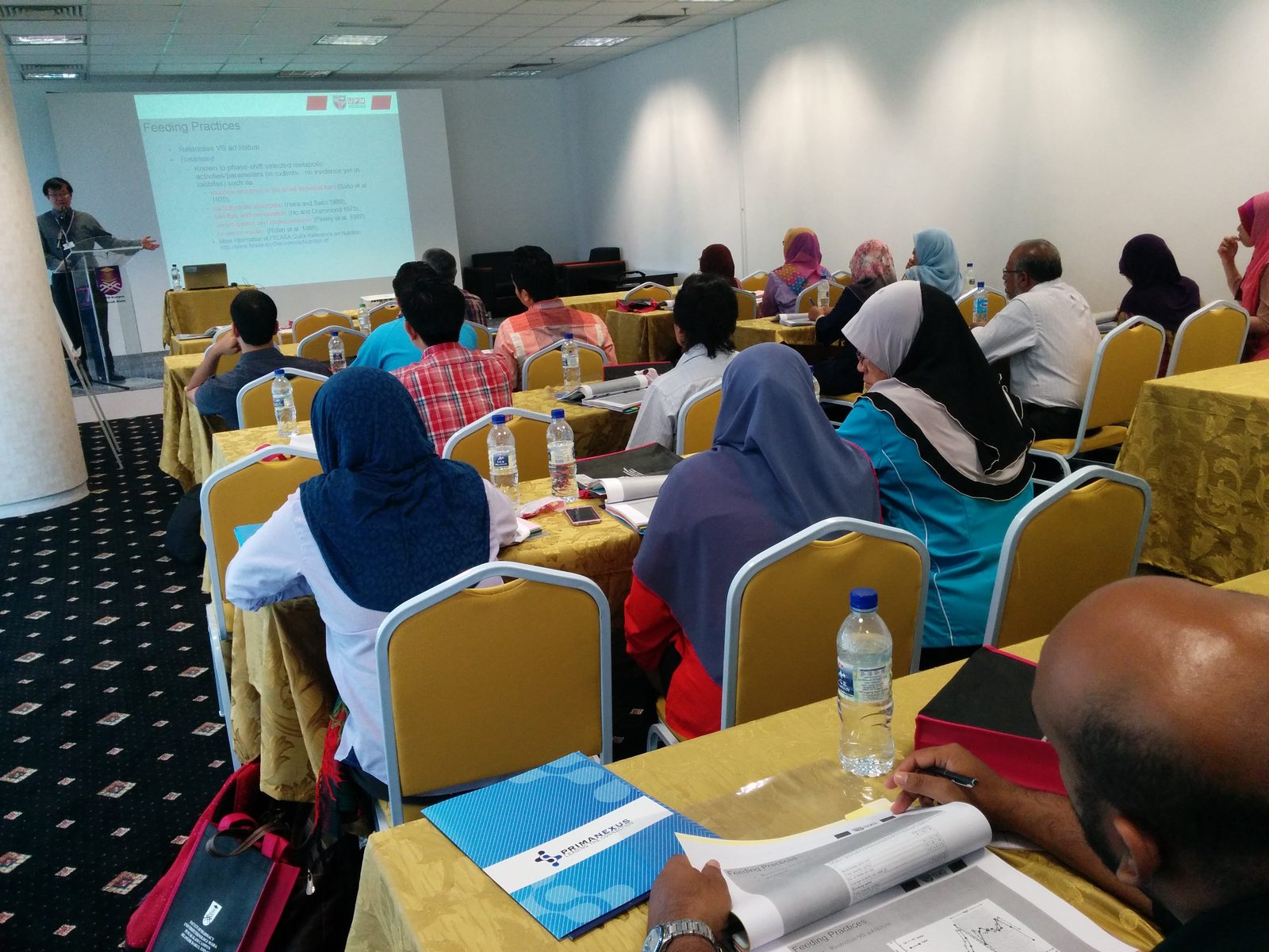 A speaker at the front of the lecture room; participants at tables, with a Prima Nexus folder on the nearest table