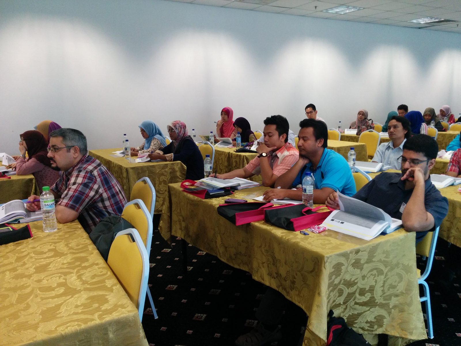 Participants seated at long tables follow a talk with printed notes and folders open in front of them