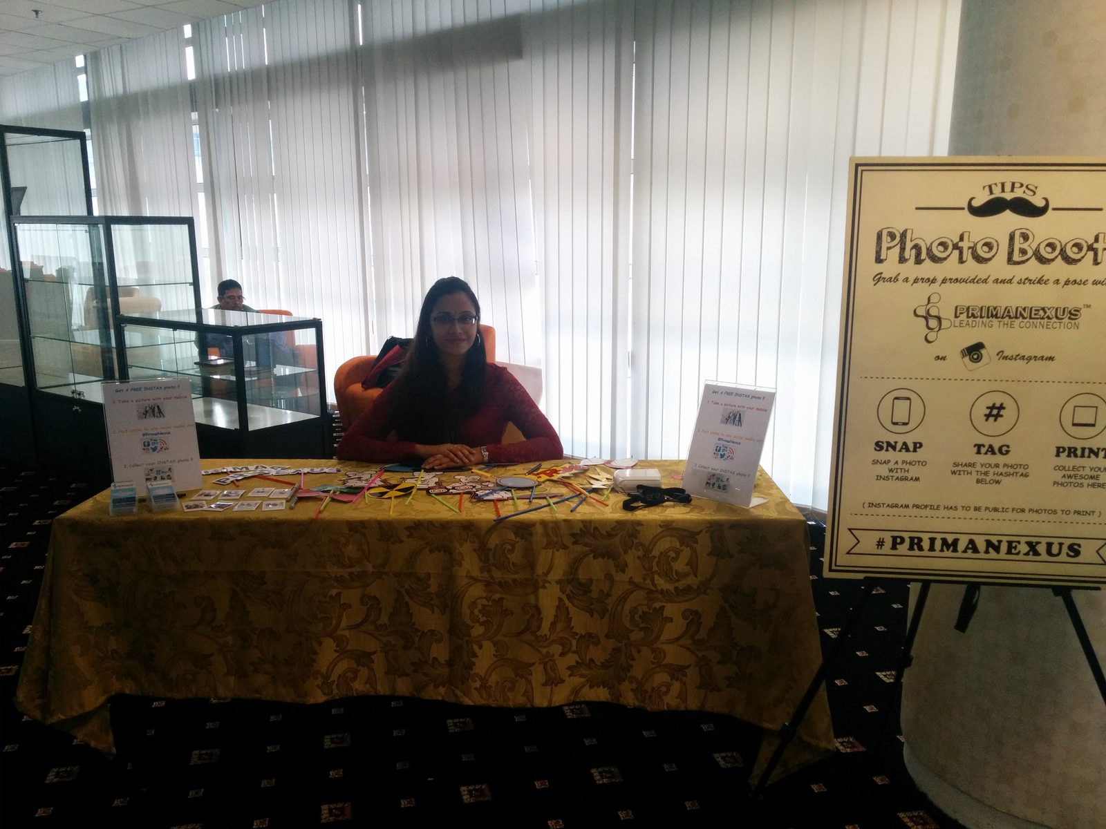 A woman seated at the Prima Nexus photo booth table with props laid out, beside a sign reading snap, tag, print