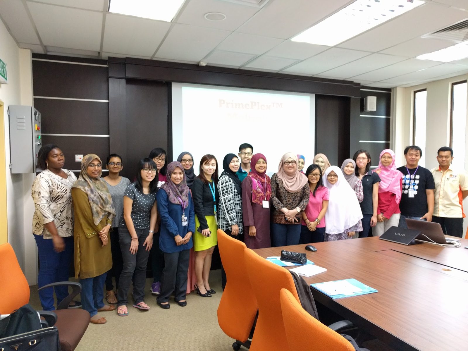 Participants and trainers stand for a group photo in the seminar room, with a title slide on the screen behind them
