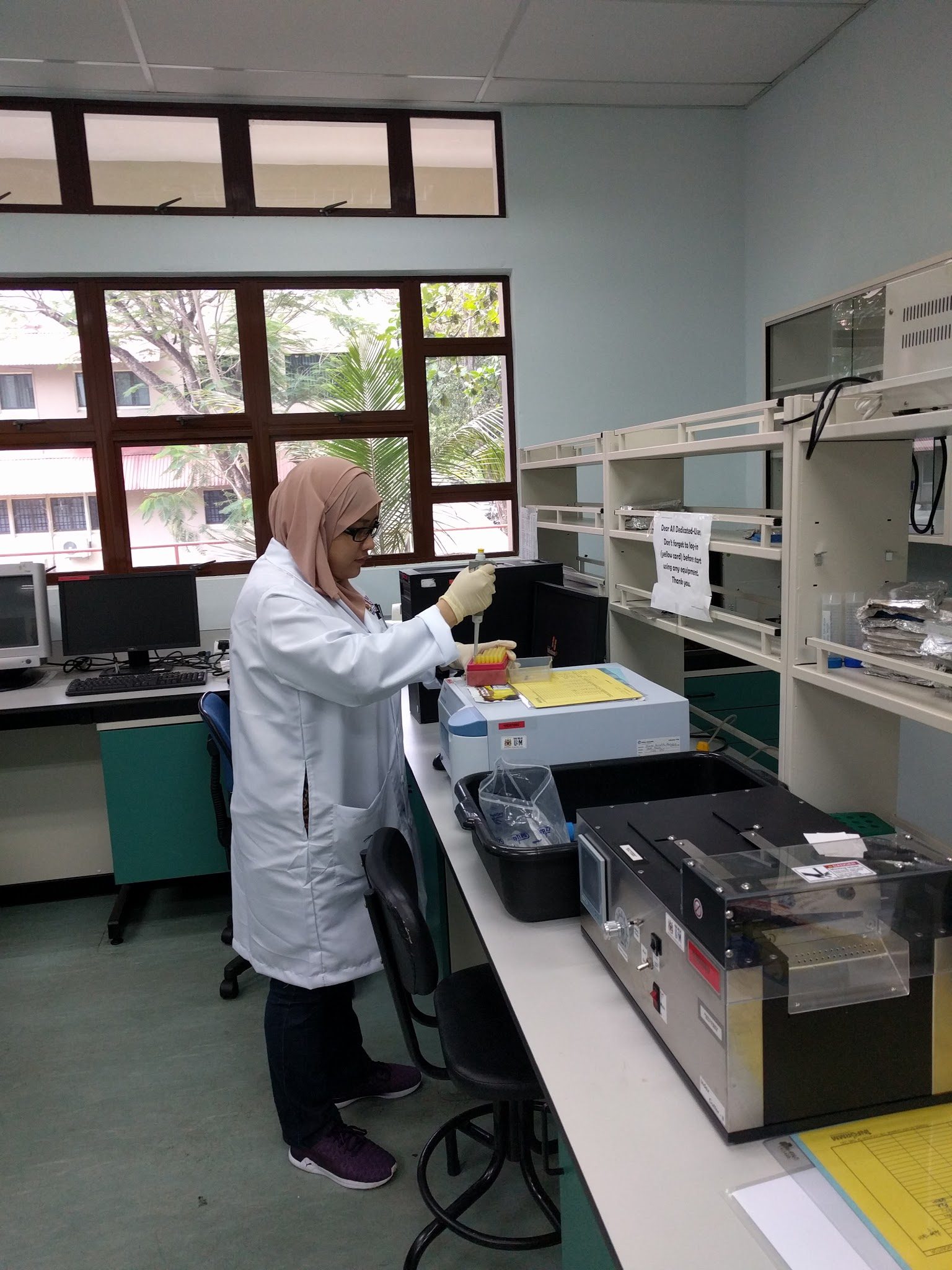A woman in a lab coat and gloves pipettes at a bench beside laboratory instruments