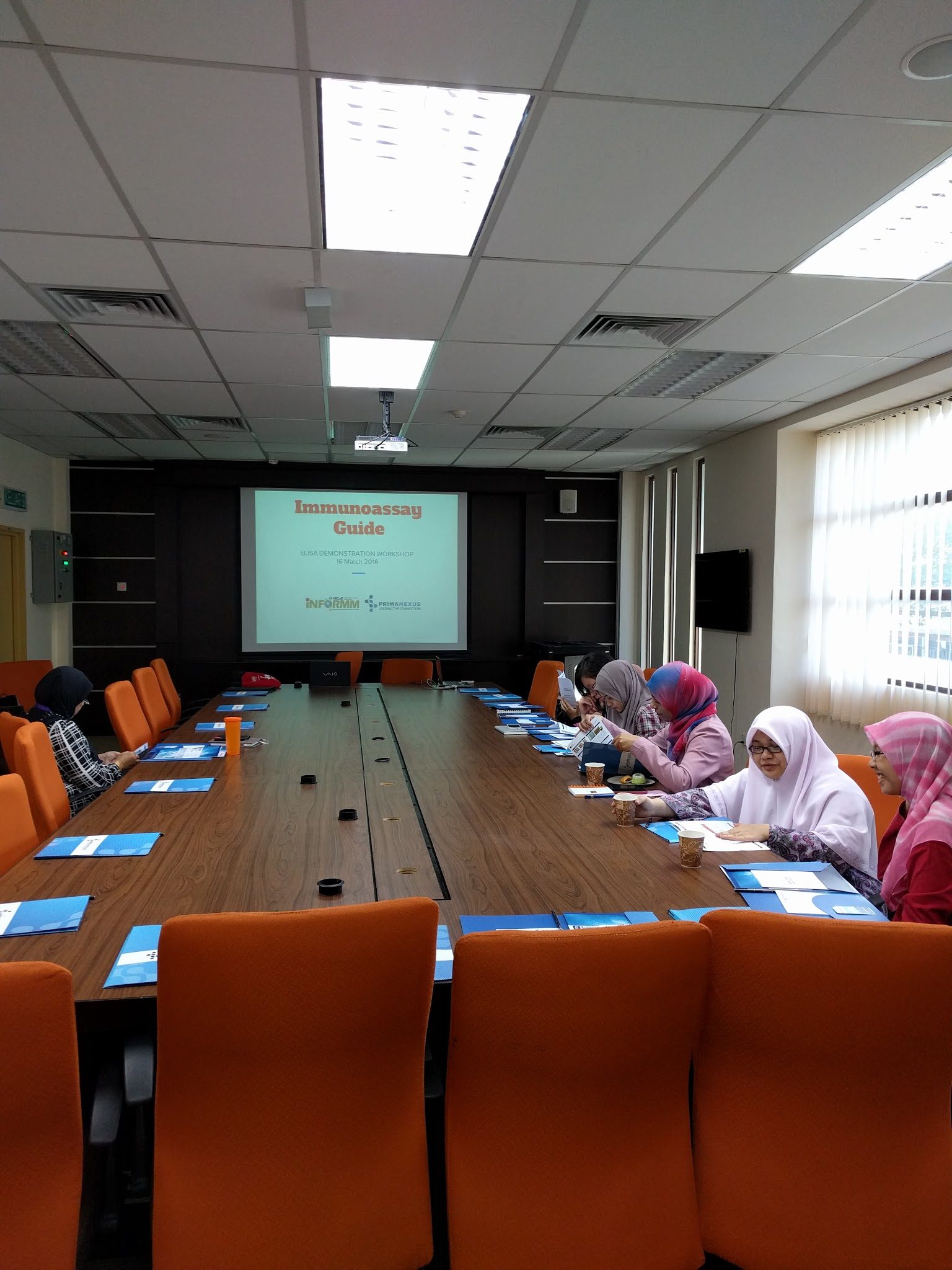 Participants at a long table read their handouts, with the Immunoassay Guide title slide on the screen