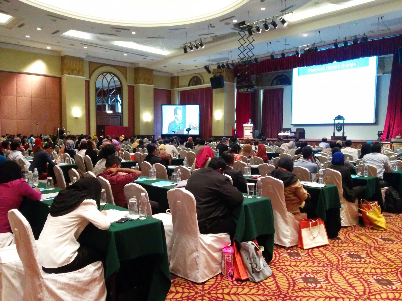 Delegates seated at long tables in a hotel hall, facing a speaker at the lectern and two large screens