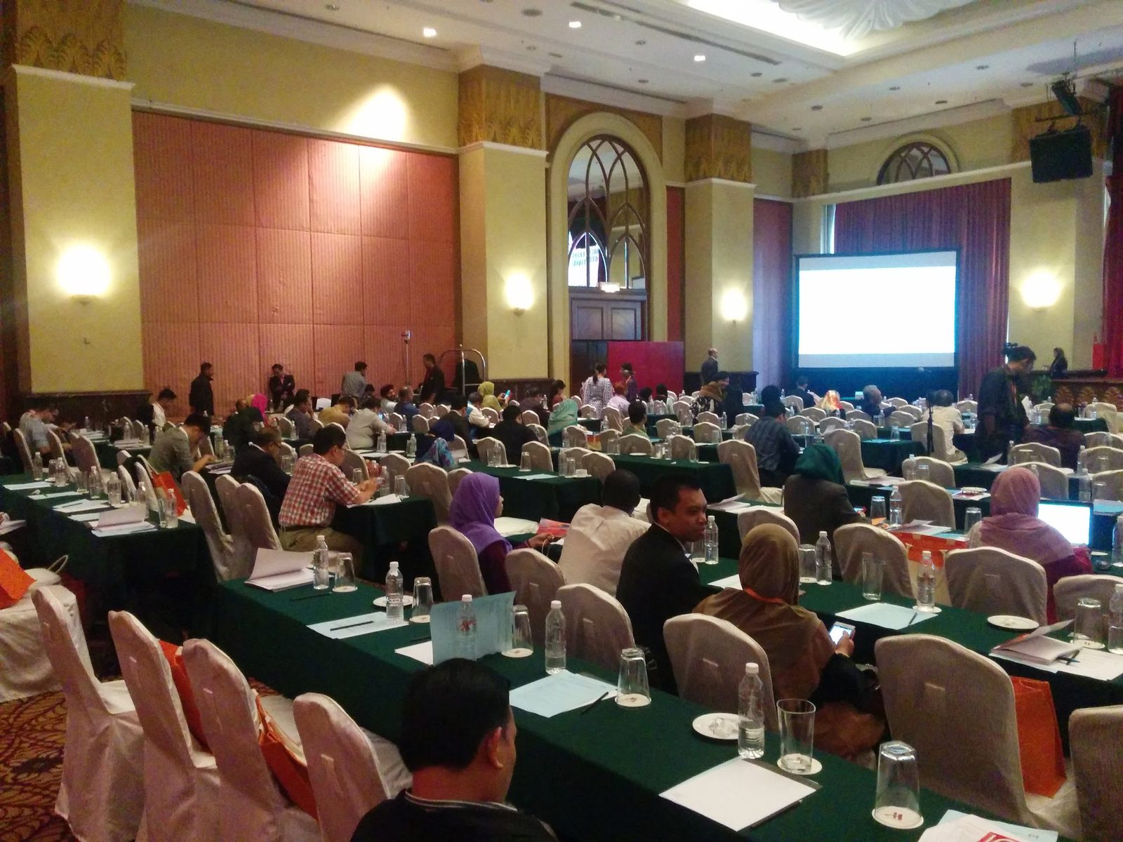 A full conference hall with delegates at long tables facing a projection screen