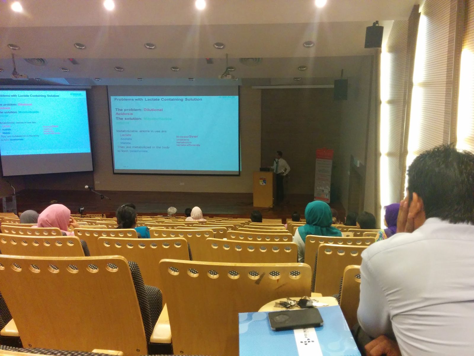 Audience in a lecture hall watching a talk, with slides on two screens and a speaker at the lectern