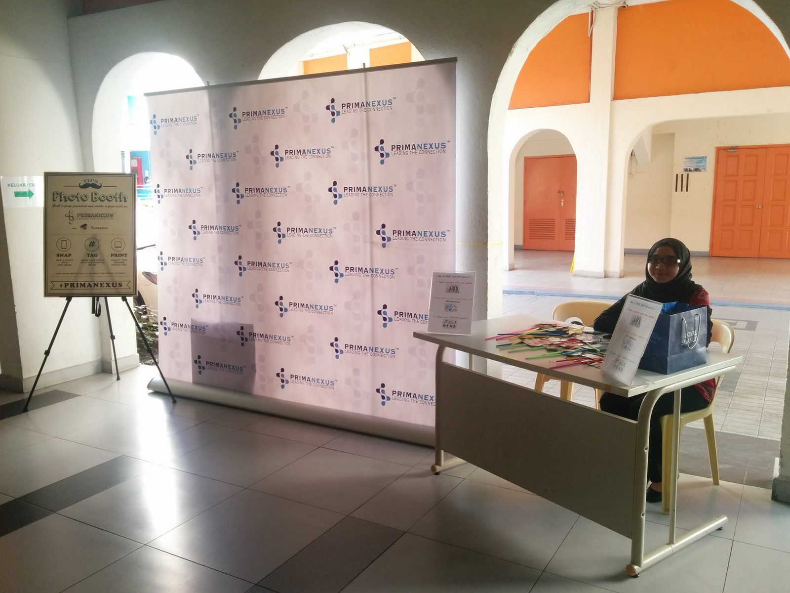 Prima Nexus photo booth: a logo backdrop, a Photo Booth sign on an easel and a props table with a person seated at it