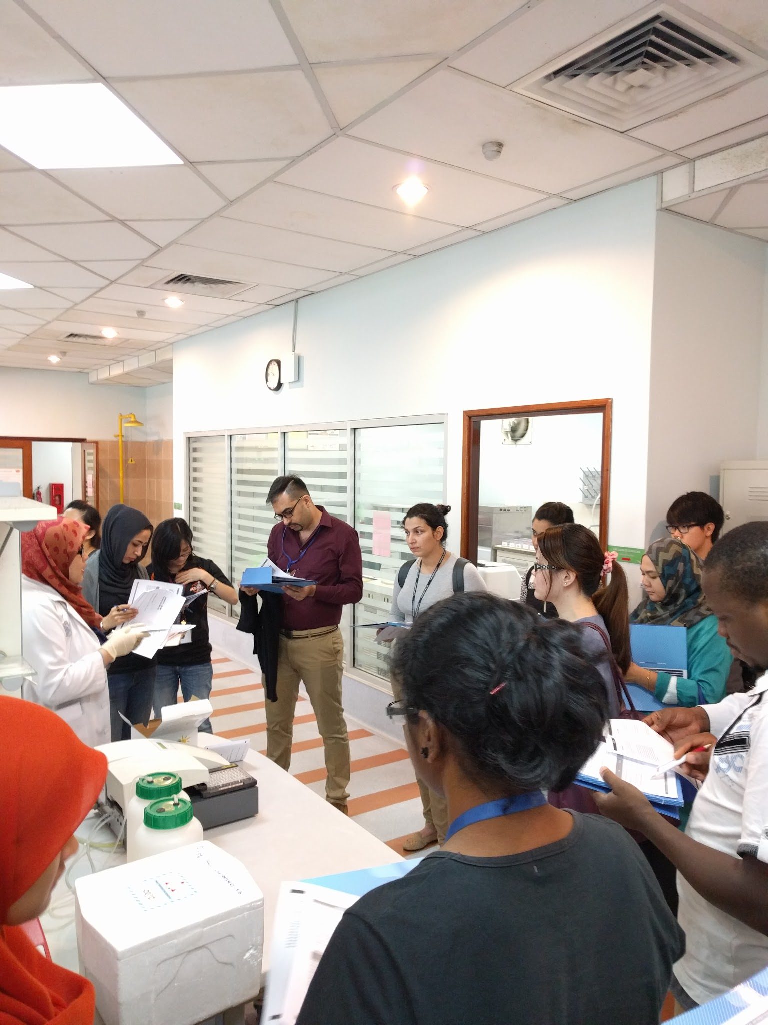 A group of participants with printed notes and folders gathers around a laboratory bench