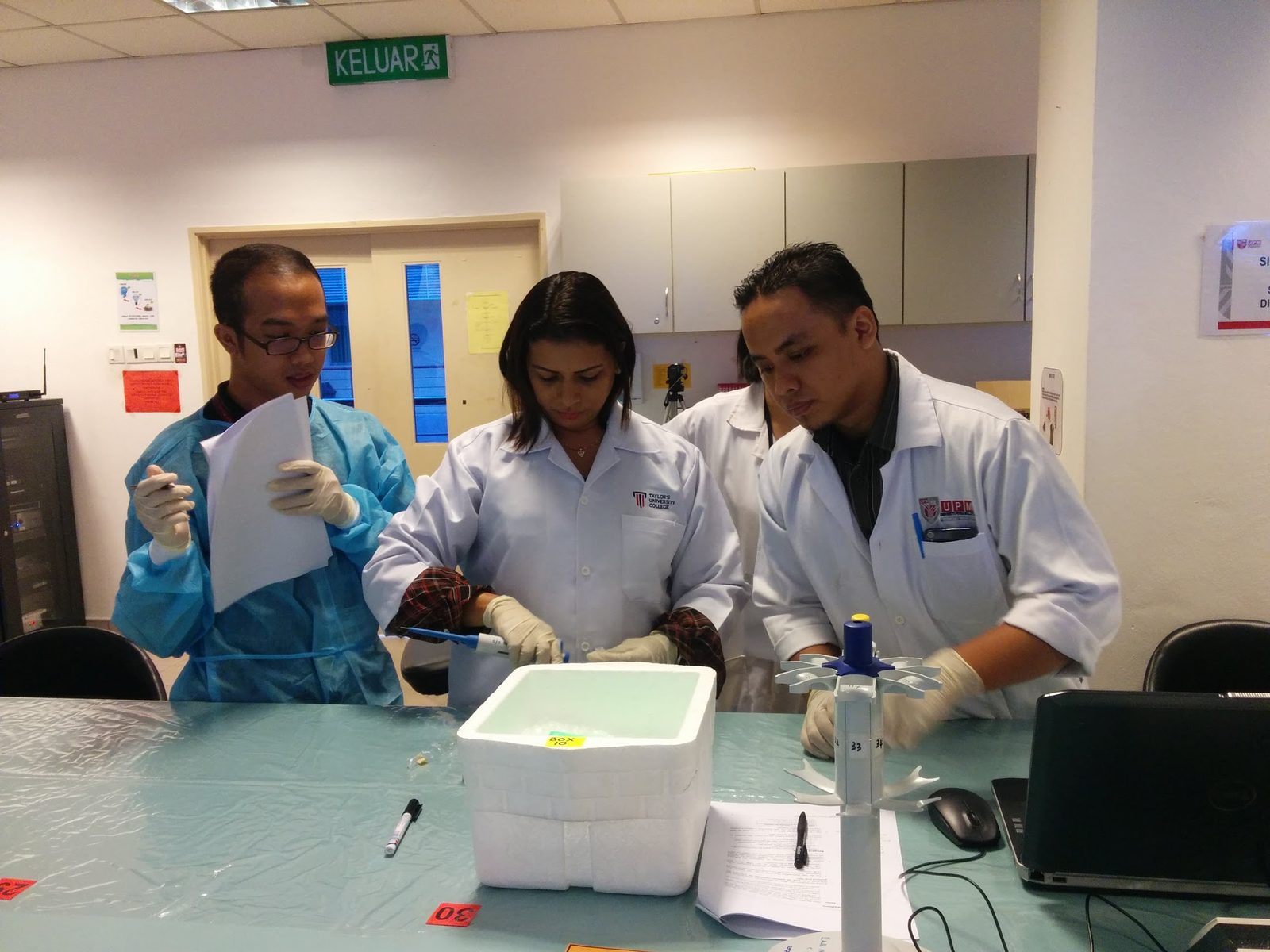 Three people in lab coats, gowns and gloves working at a laboratory bench with a sample box