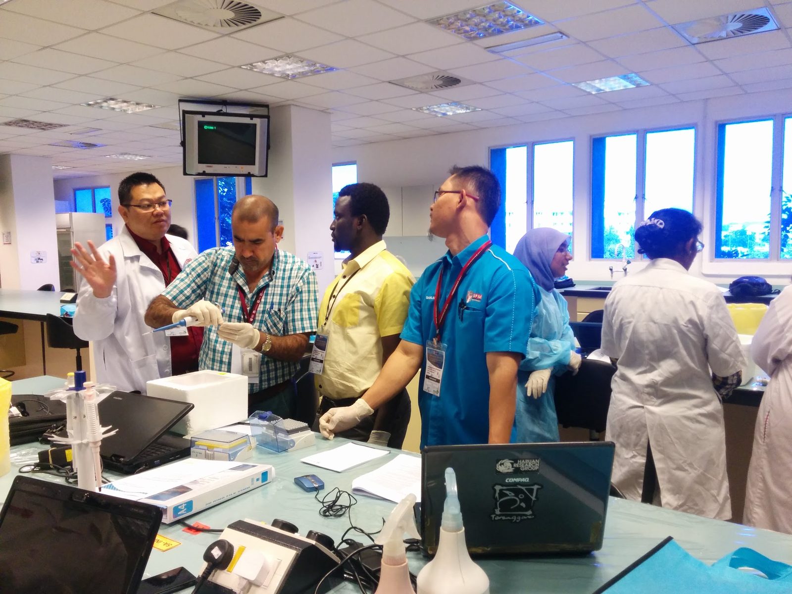 A man in a lab coat explains a step to a group standing at a laboratory bench