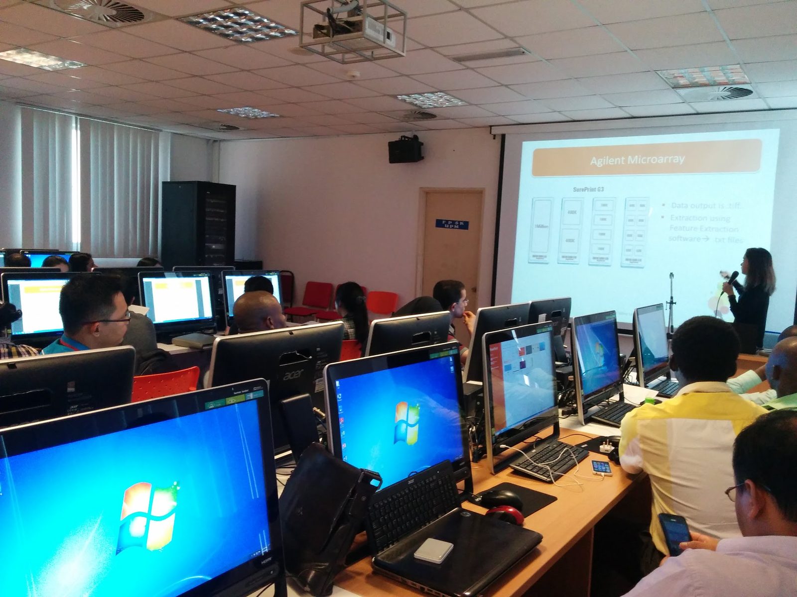 Participants seated at computers in a computer room, with a presenter and a slide at the front