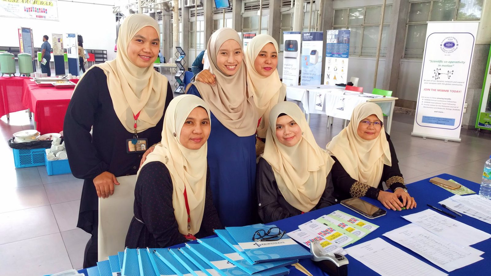 Six women at a registration desk with a stack of blue Prima Nexus folders on the table in front of them
