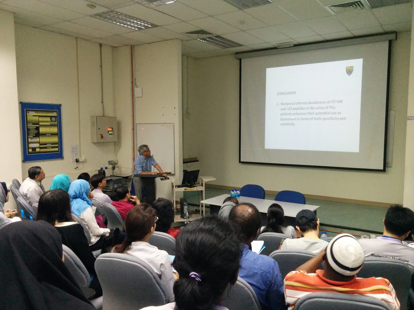 A speaker at the lectern presents a slide to a seated audience in a lecture room
