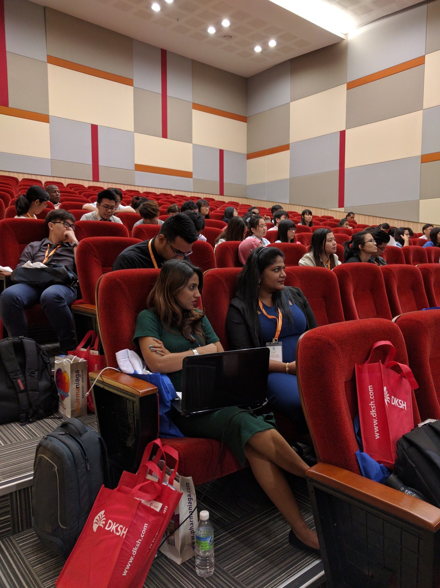 Delegates seated in rows of red seats in the auditorium, some with laptops and conference bags.