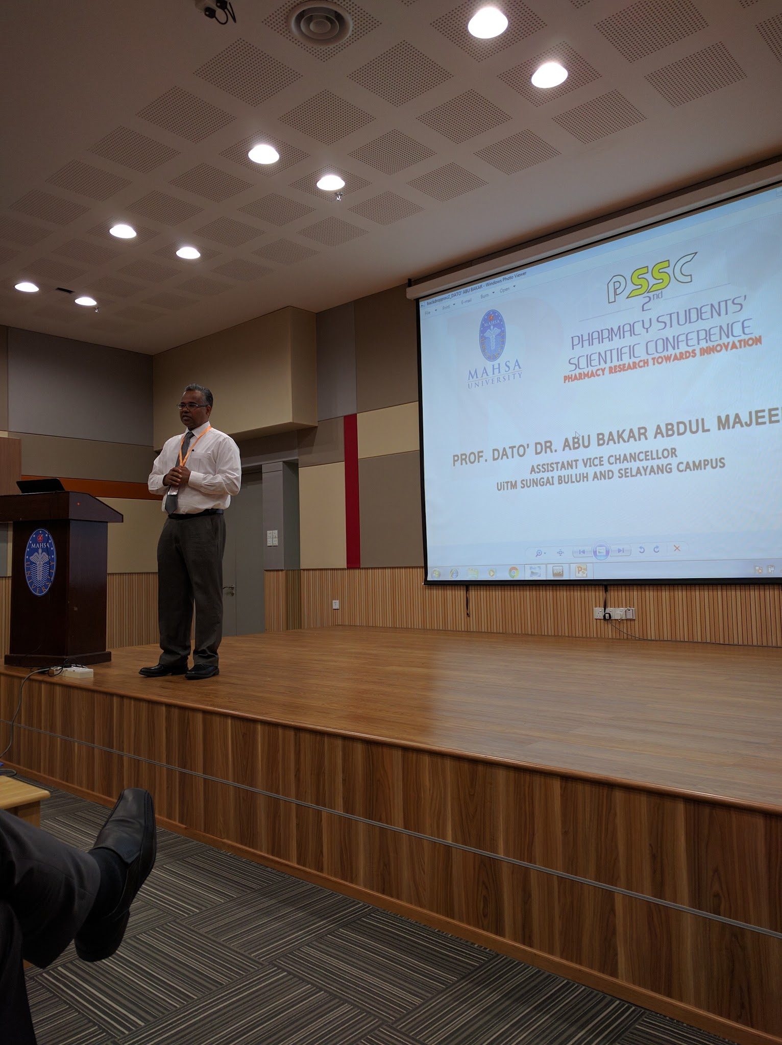 A speaker on stage addresses the audience, with the conference slide on the screen behind him.