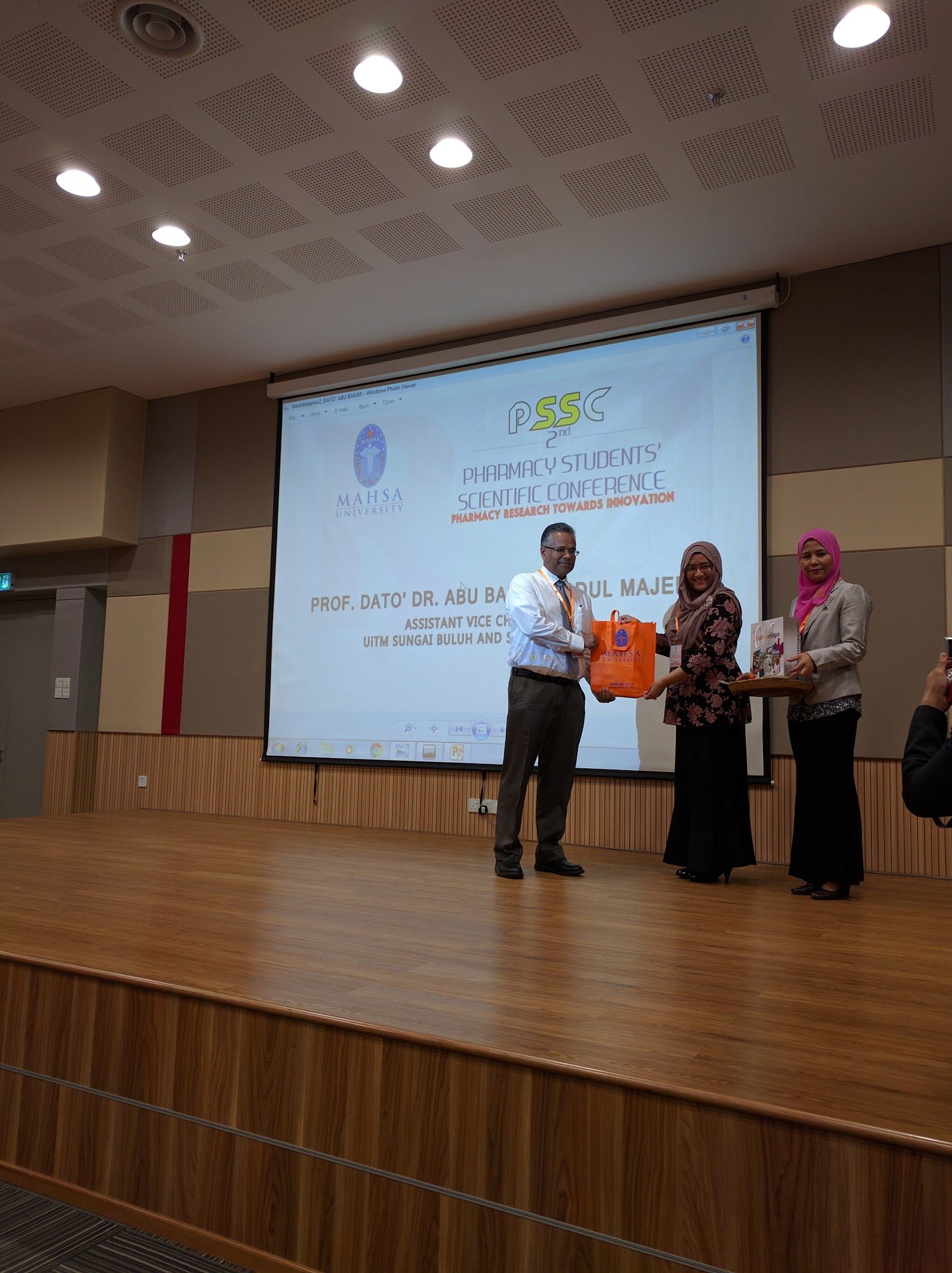 Three people on stage hand over a gift bag in front of the conference screen.