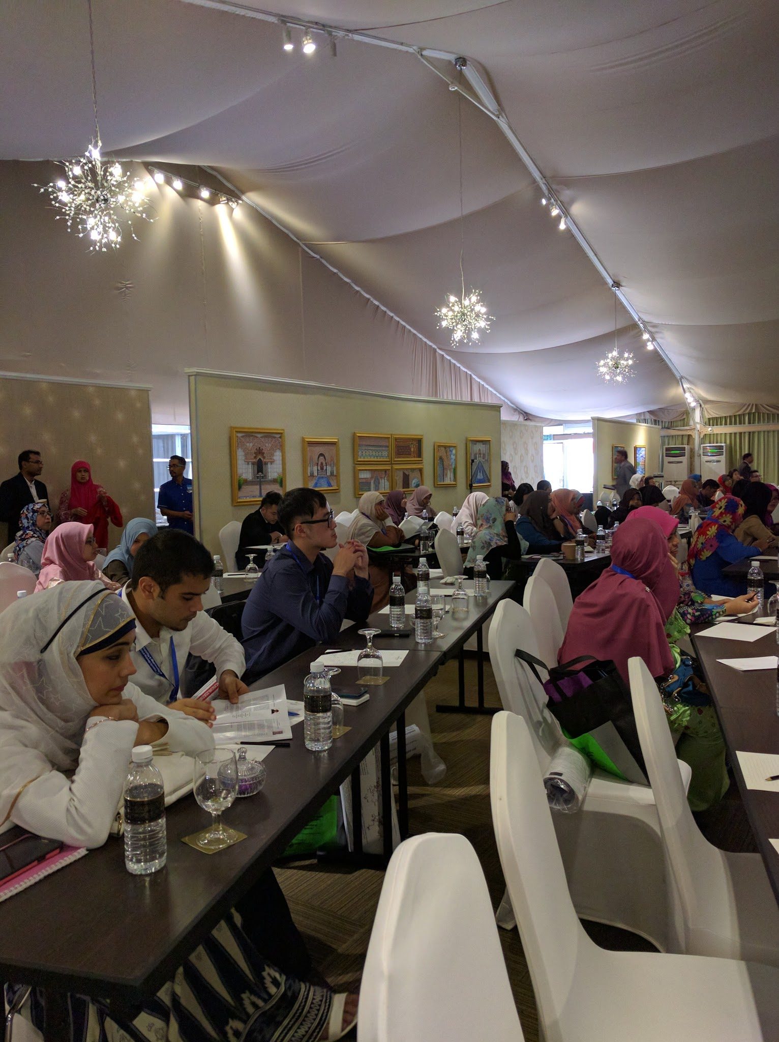 Delegates seated at long tables under a draped ceiling with pendant lights during a session