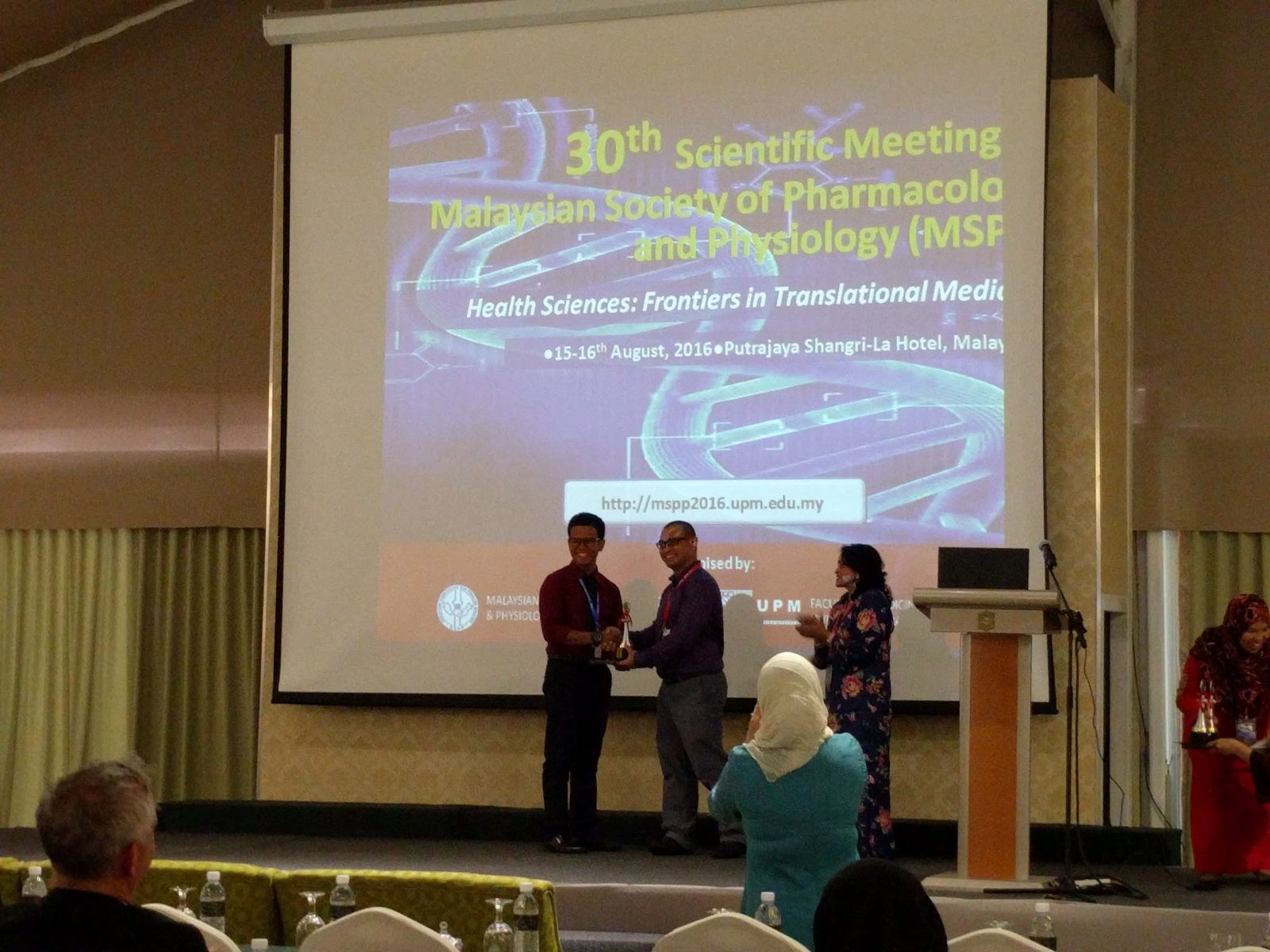 A trophy handed over on stage in front of the MSPP 2016 screen as the audience watches