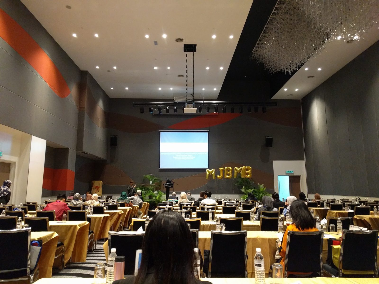 Delegates seated at long tables in the conference hall, facing a screen and a stage with MSBMB balloon letters