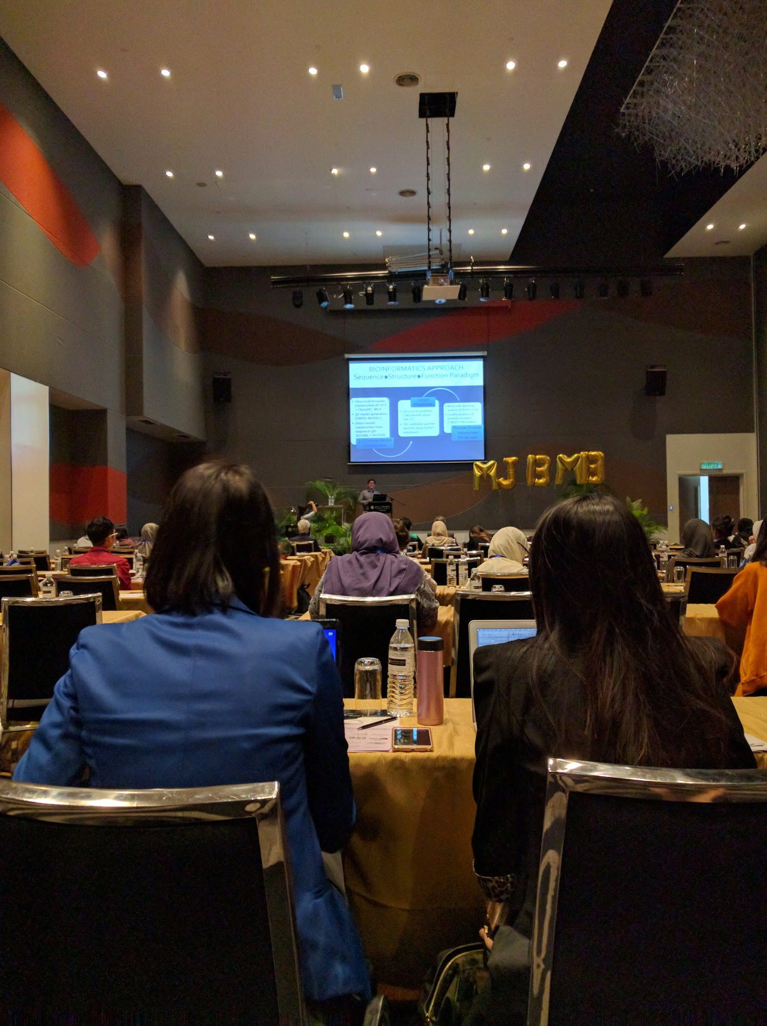 Delegates seen from behind in the hall, with a bioinformatics slide on the screen and a speaker at the lectern