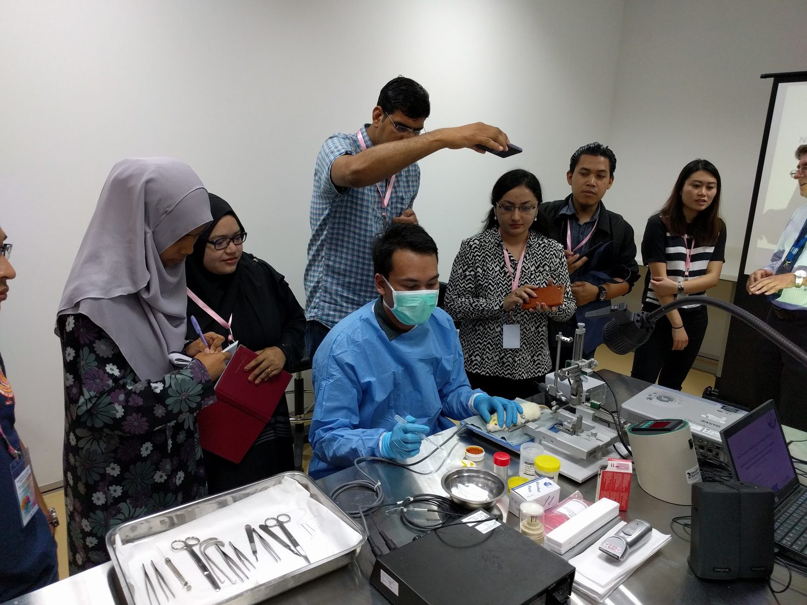Participants watch a gowned demonstrator at a stereotaxic frame, with surgical instruments laid out on the bench