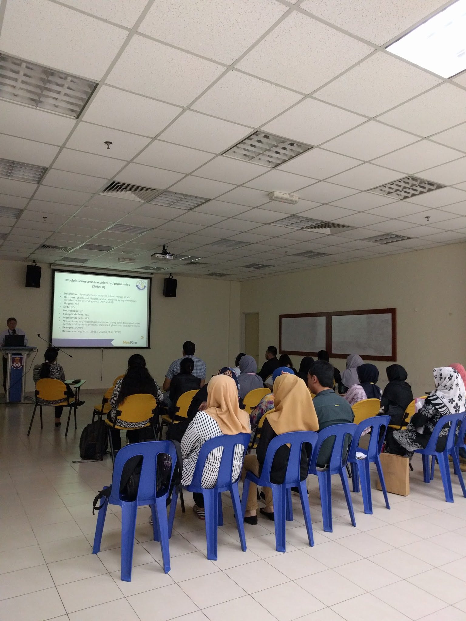 Participants seated in a seminar room listen to a speaker at the lectern, with a slide on mouse models