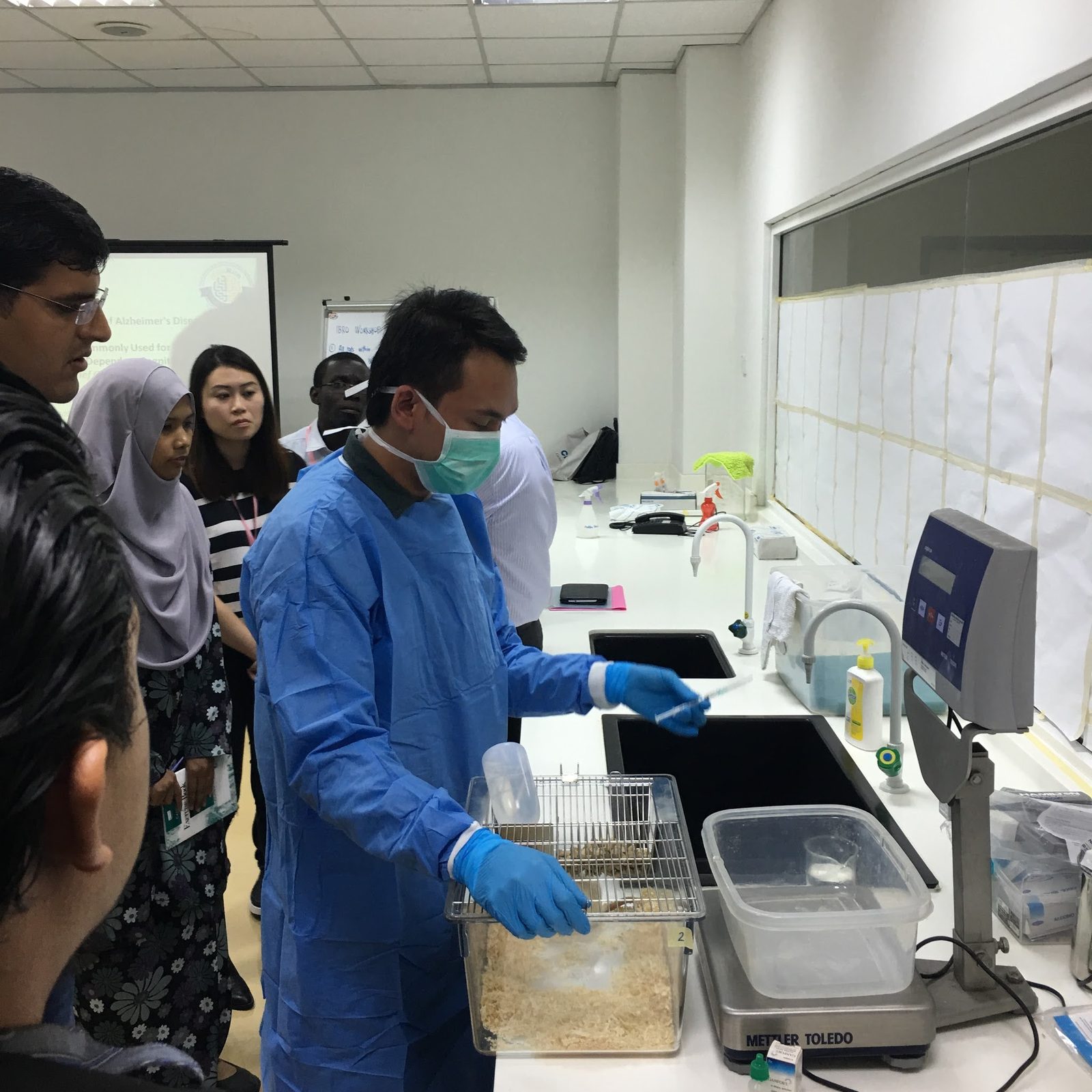 A demonstrator in a blue gown handles a clear rat cage beside a laboratory balance as participants look on