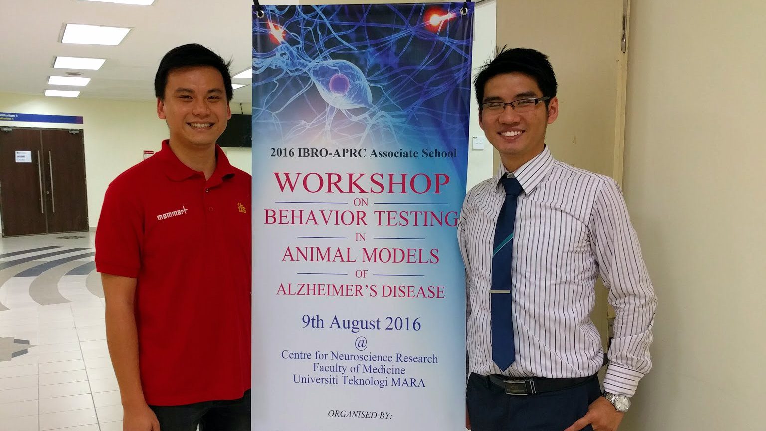 Two men stand beside a tall banner for the workshop on behaviour testing in animal models of Alzheimer's disease