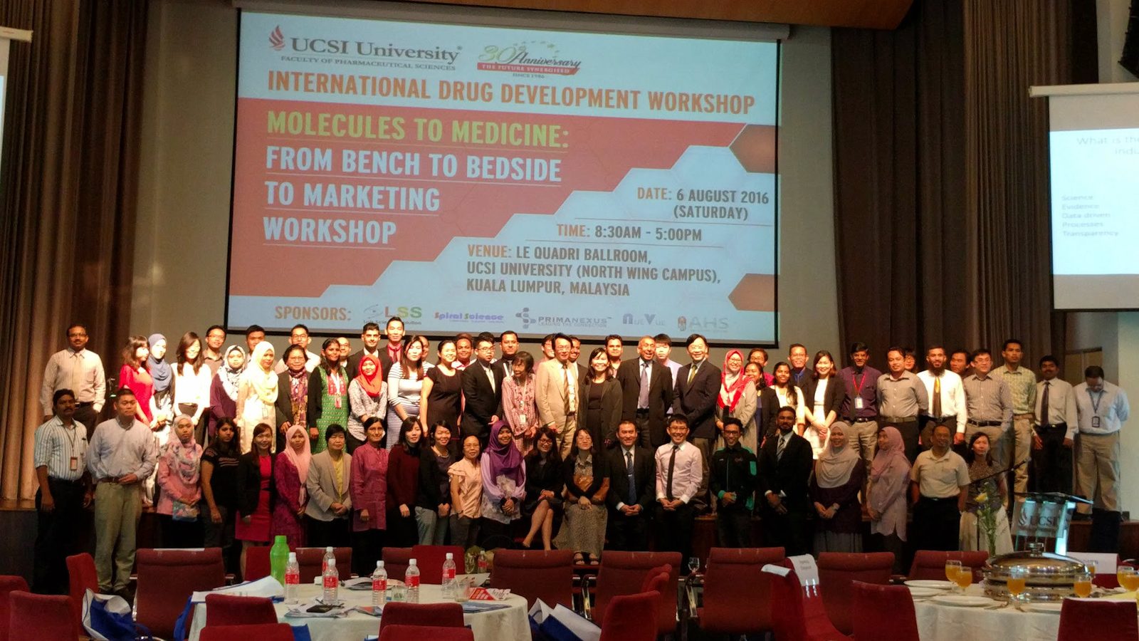 Large group of workshop participants on stage in front of a screen showing the workshop title and sponsors