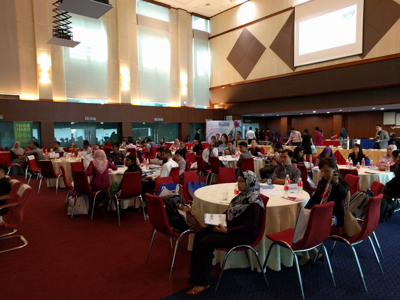 Participants seated at round tables in a large ballroom, facing a projection screen