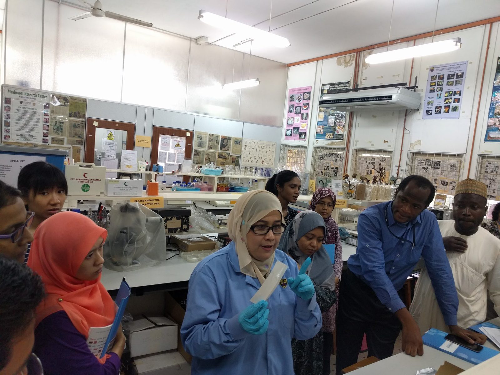A demonstrator in a blue lab coat and gloves holds up an assay item and explains it to participants at a lab bench.