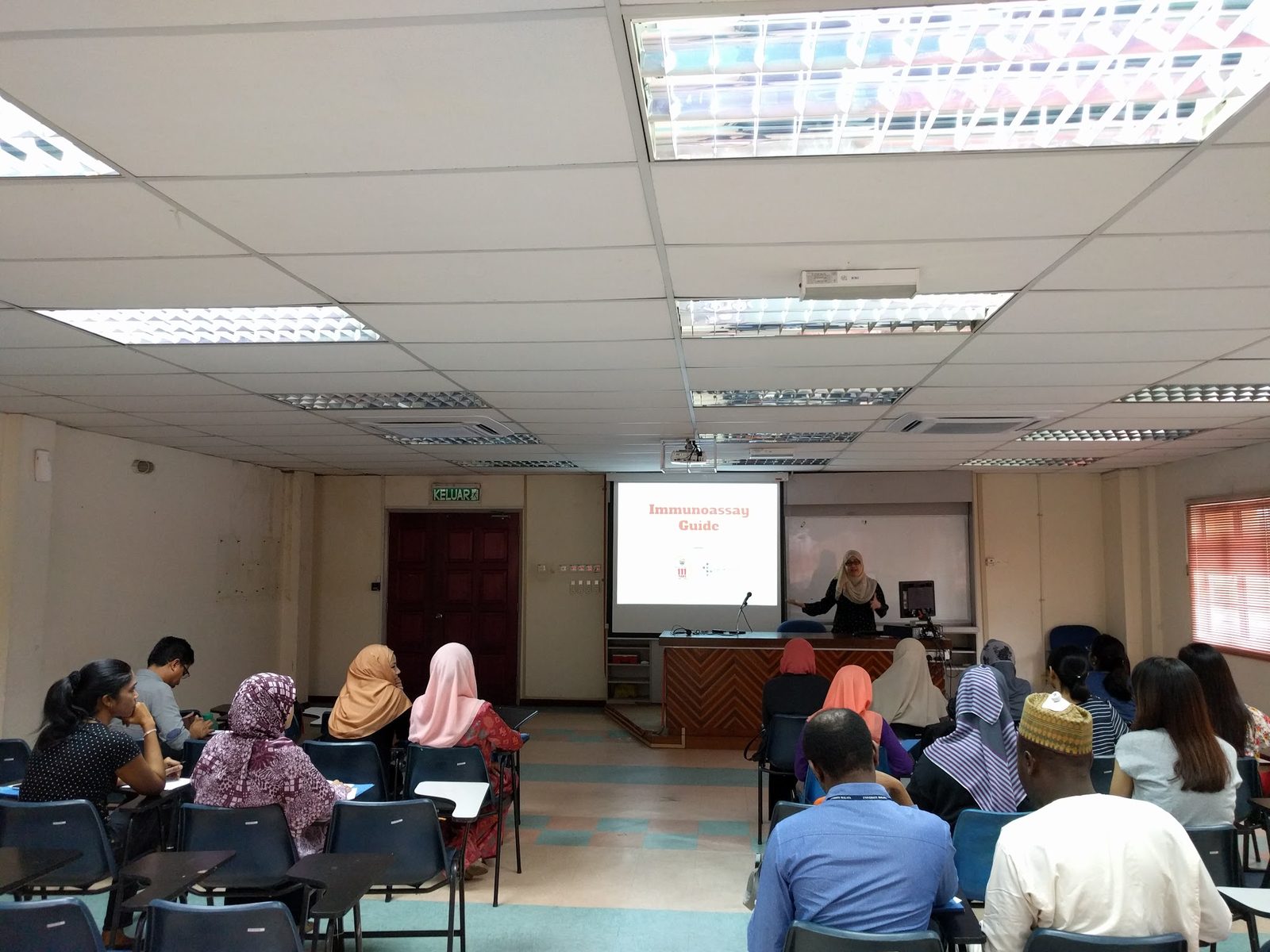 A presenter stands at the lectern beside a projected slide while participants listen in the seminar room.