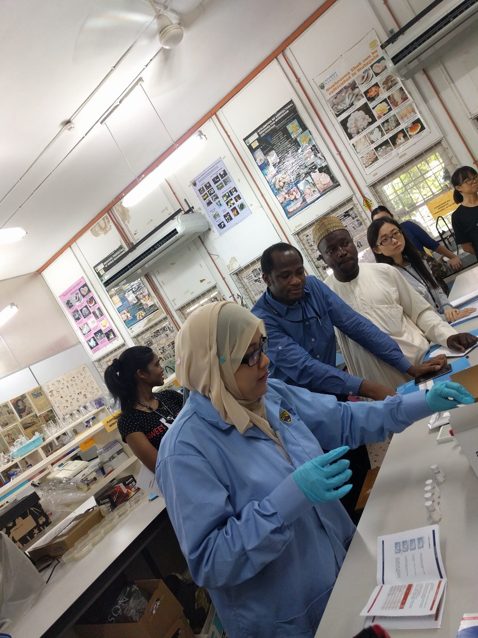 A demonstrator in a lab coat and gloves works at the bench while a participant leans in to watch.