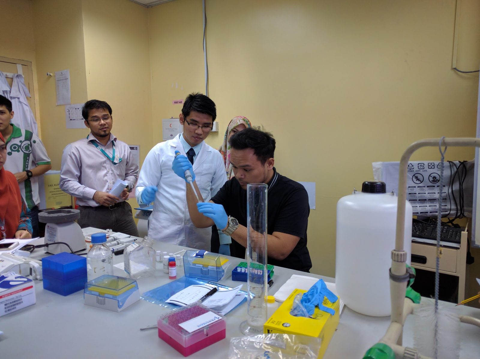 A participant and a trainer in a lab coat work with pipettes at the bench beside a tall measuring cylinder