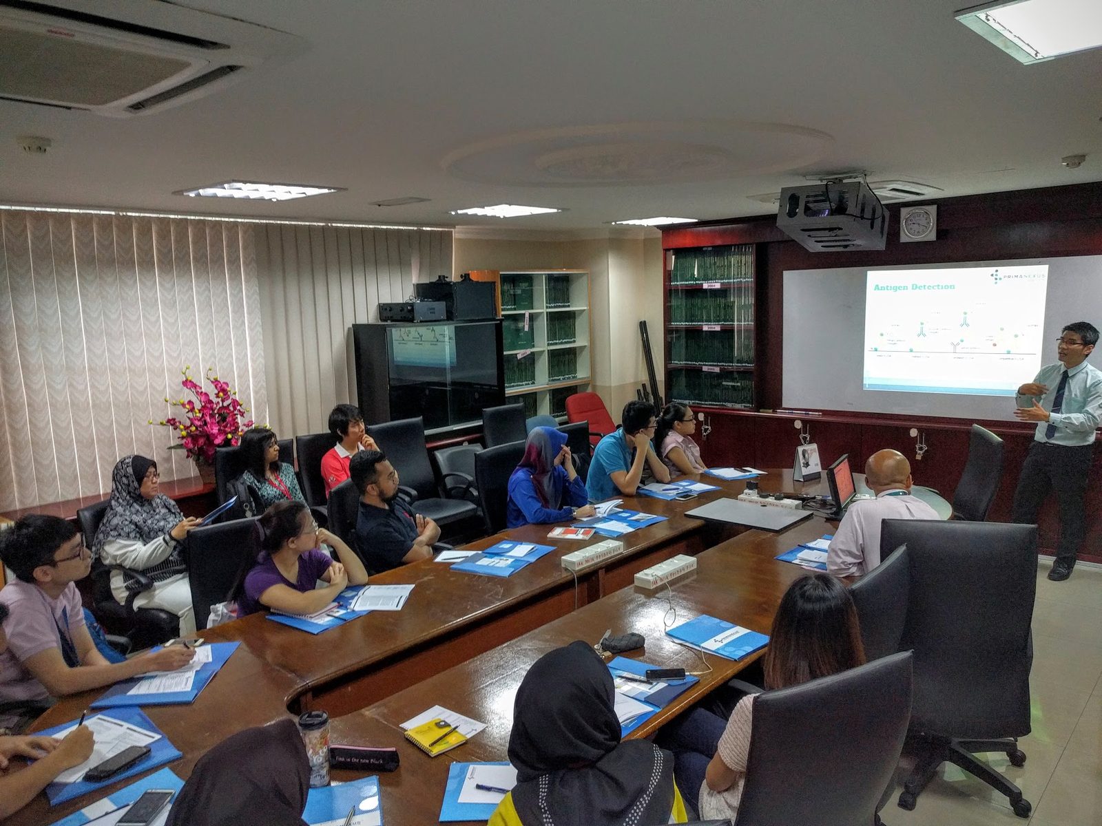 A presenter stands beside a projected slide while participants listen around a long meeting table