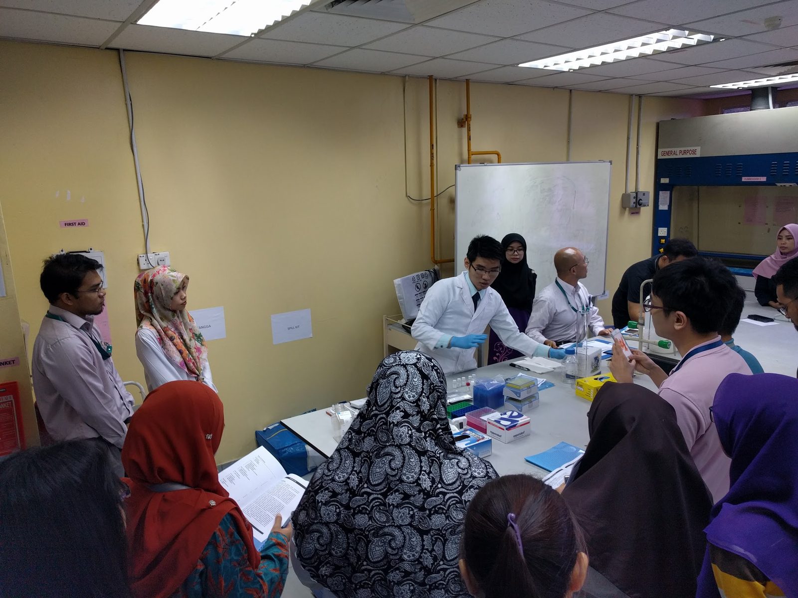 Participants gather around a lab bench to watch a trainer in a lab coat demonstrate an ELISA step
