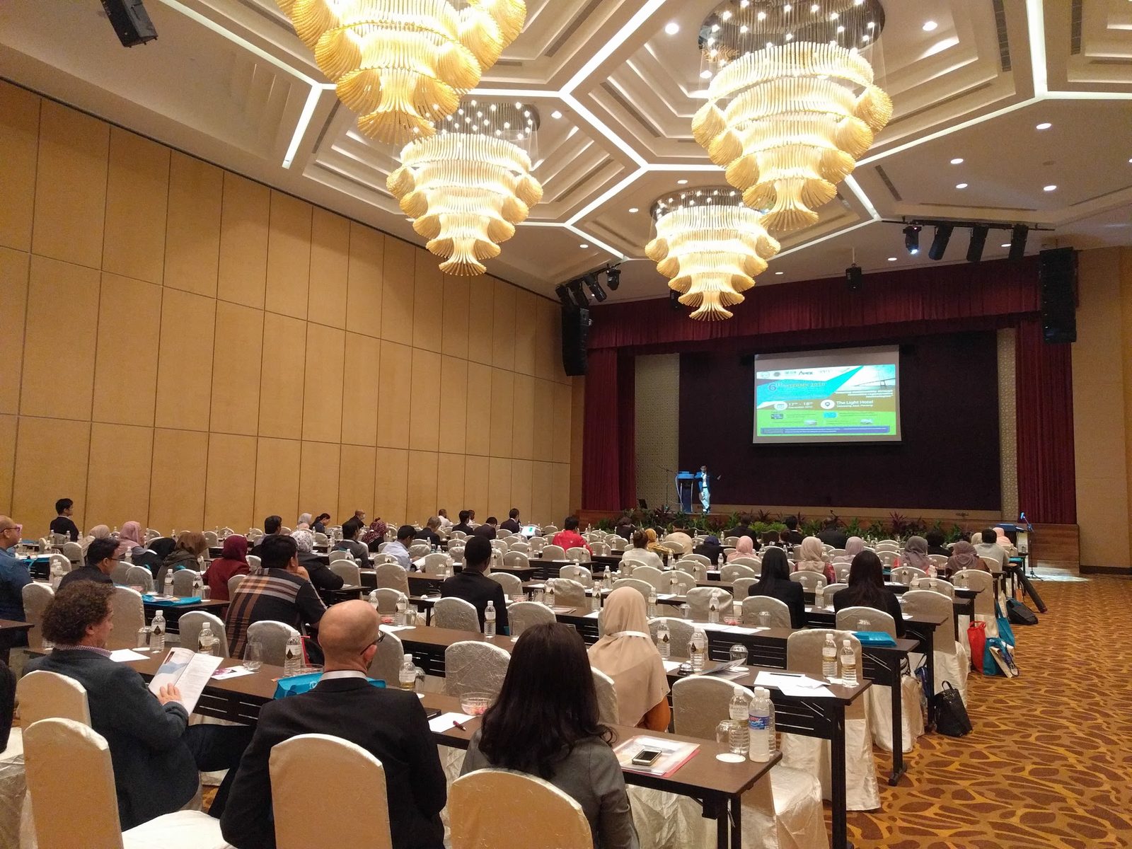 Delegates seated at long tables in a large hotel ballroom, facing the stage and a screen