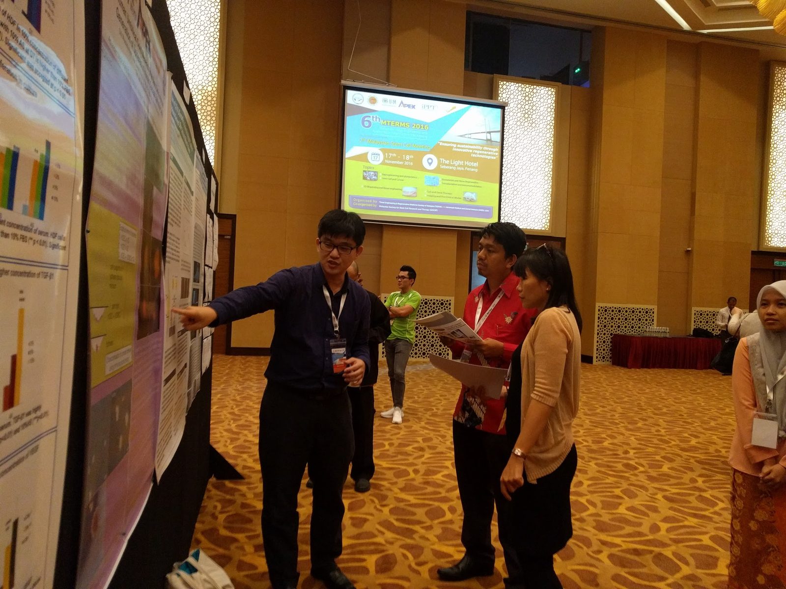 A man points to a research poster as two visitors listen, with the meeting slide on a screen behind