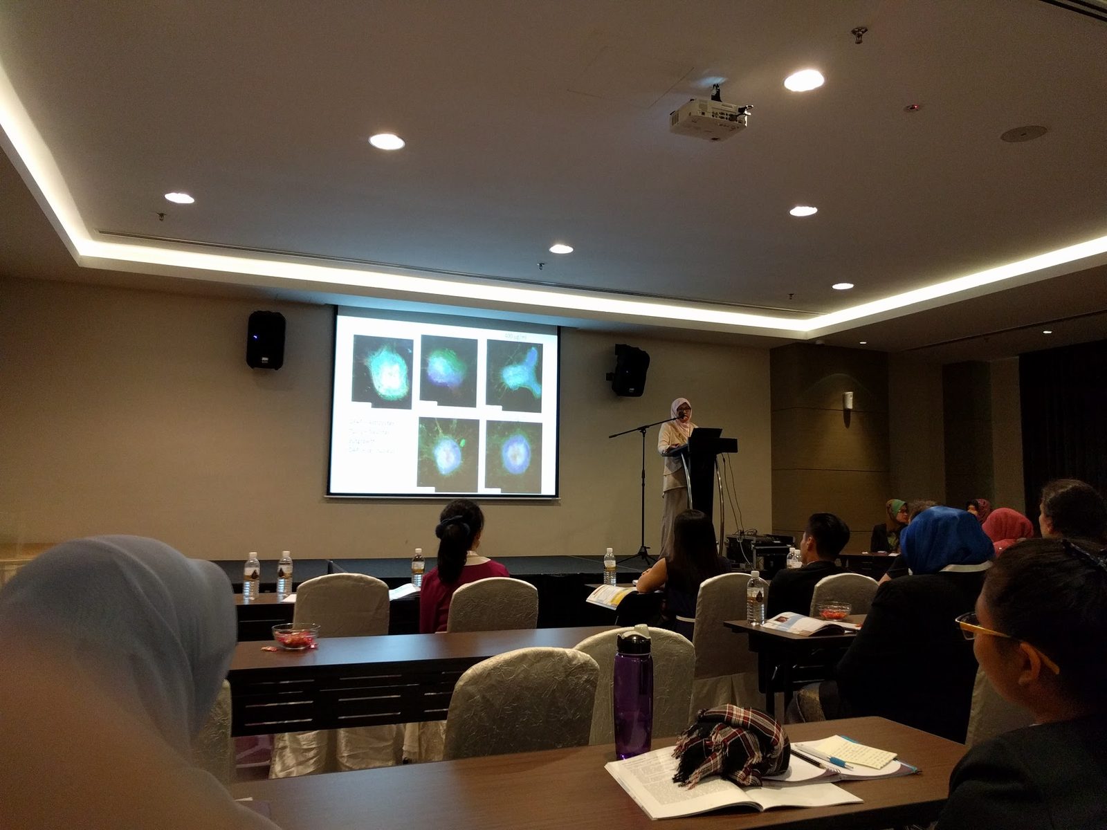 A speaker at a lectern presents microscope images of cells to a seated audience in a smaller room