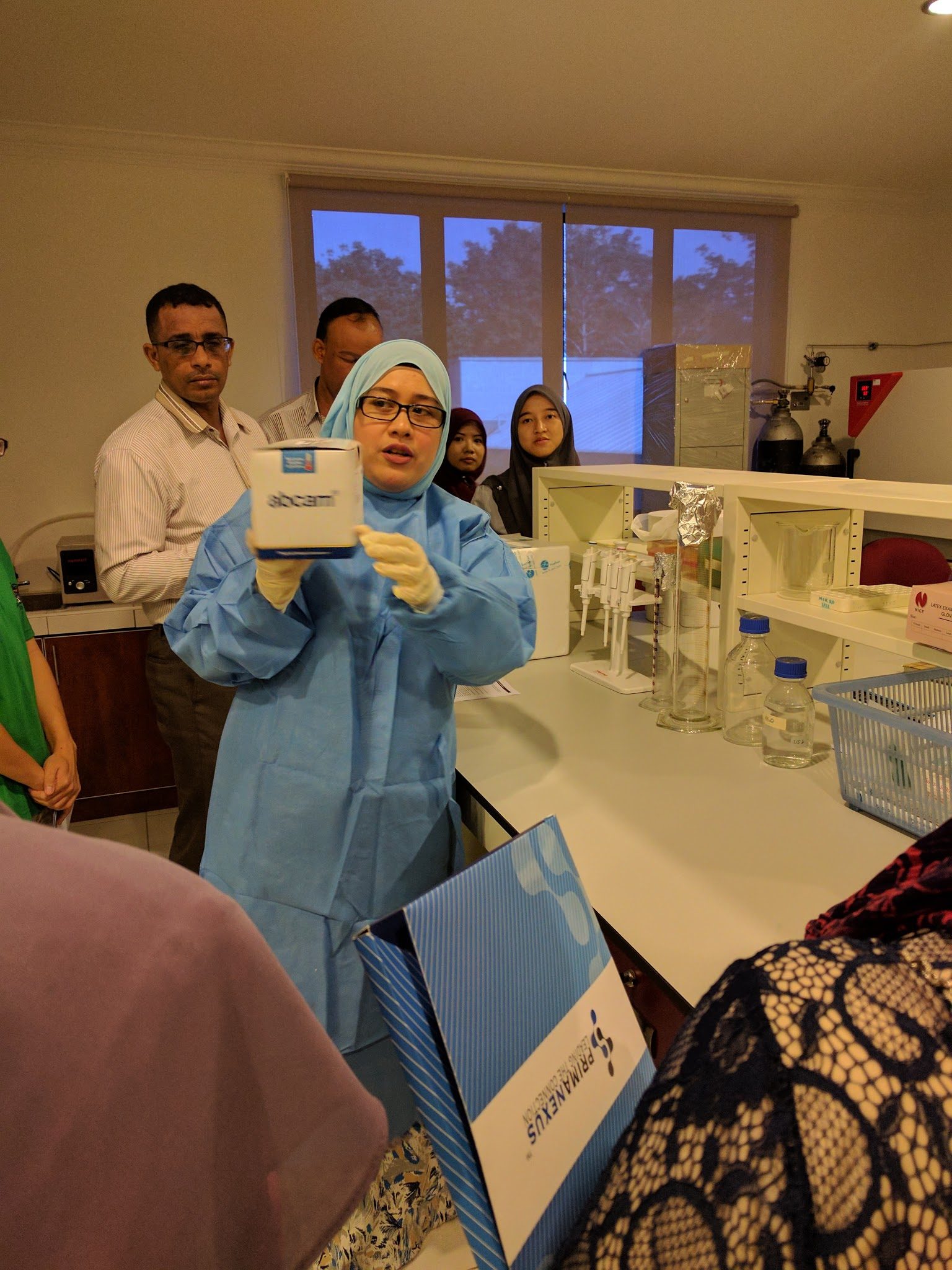 A trainer in a blue lab gown and gloves holds up an Abcam ELISA kit box in the laboratory