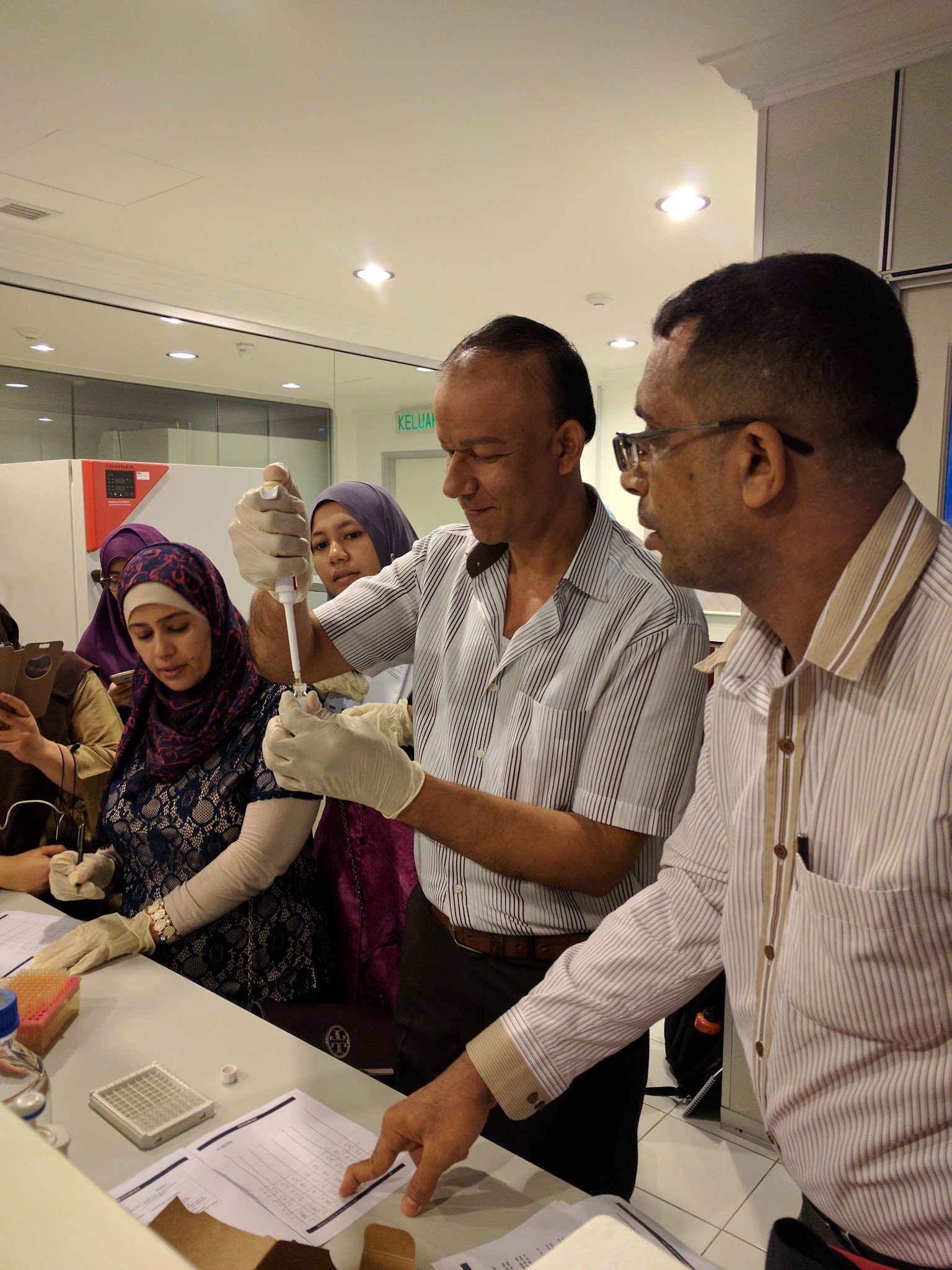 A participant wearing gloves uses a pipette at a laboratory bench while another holds the tube and others watch